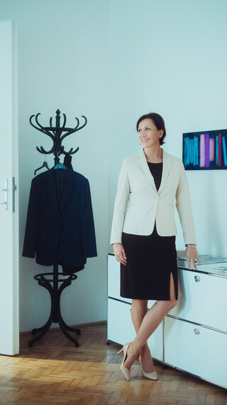 Eine Frau im schwarzen Kleid und cremefarbenem Sakko legt ihre Hand auf eine Bürokommode. Im Hintergrund sind eine Garderobe und ein buntes Bild an der Wand zu sehen.