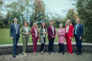 Acht Personen in Anzügen verschiedener Farben stehen draußen, mit einer Wiese und Bäumen im Hintergrund.
