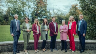 Acht Personen in Anzügen verschiedener Farben stehen draußen, mit einer Wiese und Bäumen im Hintergrund.