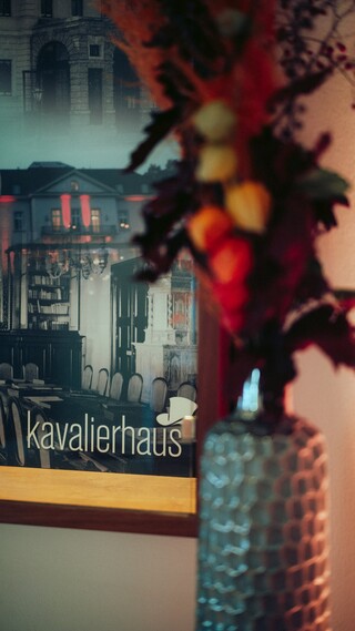 Ein Blumengesteck steht vor einer Fotografie mit der Aufschrift "kavalierhaus".