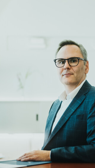 Ein stehender Herr in Anzug hat seinen Arm auf einem Tisch abgelegt.