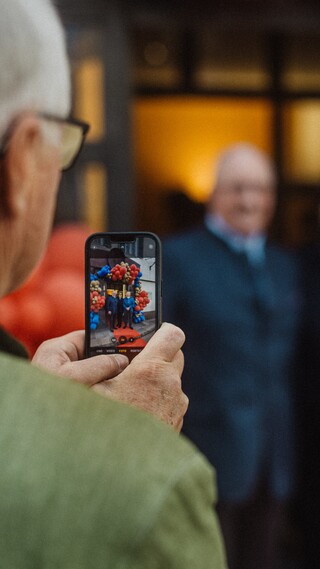 Blick über die Schulter eines älteren Herren, der mit dem Smartphone drei festlich gekleidete Personen fotografiert.