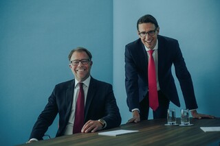 Zwei lachende Herren in Anzug an einem Tisch.