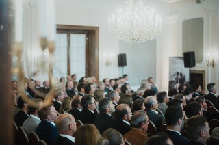 Elegant gekleidetes Publikum sitzt in einem historischen Saal unter einem Kronleuchter.