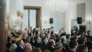 Elegant gekleidetes Publikum sitzt in einem historischen Saal unter einem Kronleuchter.