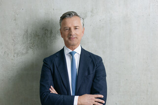 Mann mit grau-melierten Haaren und freundlichem Ausdruck steht vor einer grauen Wand. Er trägt ein weißes Hemd, eine hellblaue Krawatte und ein dunkelblaues Sakko. Er hat die Hände verschränkt.