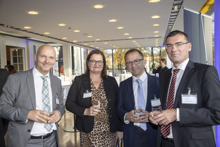 Veranstaltungsraum mit vier Personen (3 Männer, 1 Frau) im Vordergrund. Alle Personen lächeln und haben ein Glas in der Hand. Die Männer tragen dunkle Anzüge mit weißem Hemd und Krawatte, zwei Männer tragen eine Brille. Die Frau trägt ein Kleid mit braunem-Leoprint und einen schwarzen Blazer, sie trägt ebenfalls eine Brille.