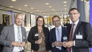 Veranstaltungsraum mit vier Personen (3 Männer, 1 Frau) im Vordergrund. Alle Personen lächeln und haben ein Glas in der Hand. Die Männer tragen dunkle Anzüge mit weißem Hemd und Krawatte, zwei Männer tragen eine Brille. Die Frau trägt ein Kleid mit braunem-Leoprint und einen schwarzen Blazer, sie trägt ebenfalls eine Brille.