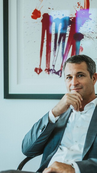 Ein Herr in Geschäftskleidung posiert sitzend vor einem Gemälde.