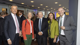 Veranstaltungsraum mit fünf Personen (3 Männer, 2 Frauen) im Vordergrund. Alle Personen lächeln. Die Männer tragen dunkle Anzüge mit weißem Hemd und Krawatte. Die eine Frau trägt einen orange-farbigen Blazer und hat gräuliche Locken, die andere trägt einen grünen Blazer und hat lange braune Haare.