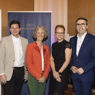 Mehrere Personen (4 Männer, 2 Frauen mittig) in einem Veranstaltungssaal, im Hintergrund ein Rollup mit dem Aufdruck "Bankhaus Spängler" ersichtlich. Alle Männer tragen dunkle Anzüge mit weißen Hemden, 1 Herr trägt ein beige-farbiges Sakko. Eine Dame trägt ein schwarzes Kleid und eine Brille, sie hat blonde hochgesteckte Haare. Die andere Frau hat gräuliche Locken und trägt einen orange-farbigen Blazer. 