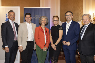 Mehrere Personen (4 Männer, 2 Frauen mittig) in einem Veranstaltungssaal, im Hintergrund ein Rollup mit dem Aufdruck "Bankhaus Spängler" ersichtlich. Alle Männer tragen dunkle Anzüge mit weißen Hemden, 1 Herr trägt ein beige-farbiges Sakko. Eine Dame trägt ein schwarzes Kleid und eine Brille, sie hat blonde hochgesteckte Haare. Die andere Frau hat gräuliche Locken und trägt einen orange-farbigen Blazer. 