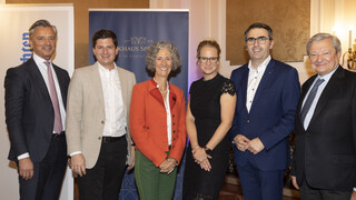 Mehrere Personen (4 Männer, 2 Frauen mittig) in einem Veranstaltungssaal, im Hintergrund ein Rollup mit dem Aufdruck "Bankhaus Spängler" ersichtlich. Alle Männer tragen dunkle Anzüge mit weißen Hemden, 1 Herr trägt ein beige-farbiges Sakko. Eine Dame trägt ein schwarzes Kleid und eine Brille, sie hat blonde hochgesteckte Haare. Die andere Frau hat gräuliche Locken und trägt einen orange-farbigen Blazer. 