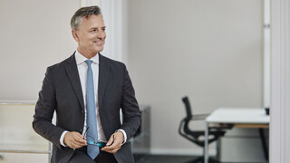 Mann im Anzug mit hellblauer Krawatte, der seine Brille in den Händen hält. Im Hintergrund befindet sich ein Schreibtisch mit Stuhl.