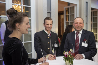 Drei Personen (2 Männer, 1 Frau) stehen bei einem Stehtisch mit weißer Husse und rotem Blumen-Arrangement. Sie halten ein Getränk mit Strohhalm und Limette in der Hand. Alle Personen lächeln. Die Frau trägt ein schwarzes Oberteil und hat hochgesteckte Haare. Ein Mann trägt einen Trachtenanzug, der andere einen Anzug mit weißem Hemd und dunkelrot-gestreifter Krawatte. 