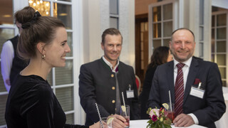 Drei Personen (2 Männer, 1 Frau) stehen bei einem Stehtisch mit weißer Husse und rotem Blumen-Arrangement. Sie halten ein Getränk mit Strohhalm und Limette in der Hand. Alle Personen lächeln. Die Frau trägt ein schwarzes Oberteil und hat hochgesteckte Haare. Ein Mann trägt einen Trachtenanzug, der andere einen Anzug mit weißem Hemd und dunkelrot-gestreifter Krawatte. 
