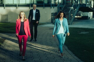 Zwei Damen und ein Herr in Geschäftskleidung gehen fröhlich auf einem gepflasterten Weg von einem Gebäude mit Wintergarten weg.