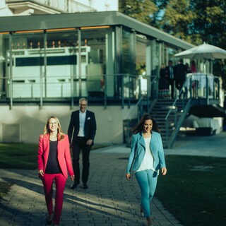 Zwei Damen und ein Herr in Geschäftskleidung gehen fröhlich auf einem gepflasterten Weg von einem Gebäude mit Wintergarten weg.