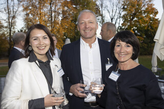 Zwei Frauen und ein Mann lächeln, alle haben ein Glas Wasser in der Hand und sind schwarz-weiß gekleidet. Sie befinden sich im Freien und im Hintergrund sind orange-leuchtende Bäume zu sehen.