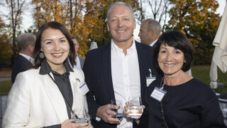 Zwei Frauen und ein Mann lächeln, alle haben ein Glas Wasser in der Hand und sind schwarz-weiß gekleidet. Sie befinden sich im Freien und im Hintergrund sind orange-leuchtende Bäume zu sehen.