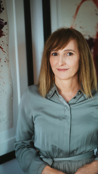 Eine Dame in Kleid posiert lächelnd vor zwei Gemälden.