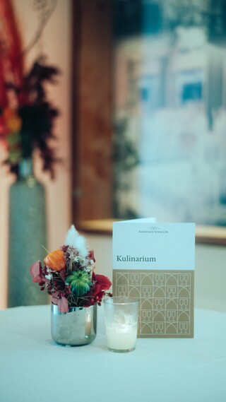 Festlich dekorierter Stehtisch vor einem Gemälde und einem Blumengesteck.