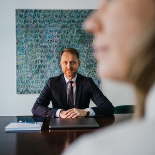 Am Tisch sitzt ein Mann im blauen Anzug, die Hände auf dem Tisch. 