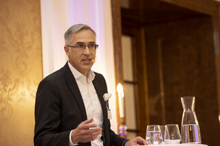 Mann mit grau-meliertem Haar und Brille spricht stehend am Podium mit einem Headset-Mikrofon. Den Hintergrund ziert eine beige-gemusterte Wand. Ebenfalls ersichtlich sind eine Karaffe Wasser und drei Gläser.