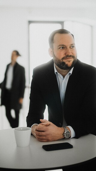 Ein Mann in schwarzem Anzug steht an einem Stehtisch und schaut nach links.