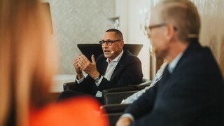 Ein sitzender Herr in Anzug mit Headset spricht und gestikuliert.