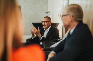 Ein sitzender Herr in Anzug mit Headset spricht und gestikuliert.