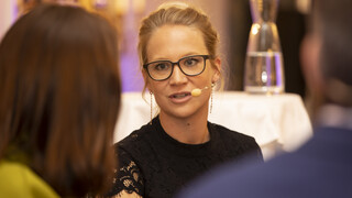 Frau mit hochgesteckten, blonden Haaren und Brille sitzt am Podium und spricht in ein Headset mit Mikrofon. Zwei weitere Personen sind von hinten leicht erkennbar.