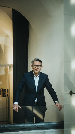 Ein lächelnder Herr in Anzug stützt sich mit den Händen auf ein gläsernes Geländer.