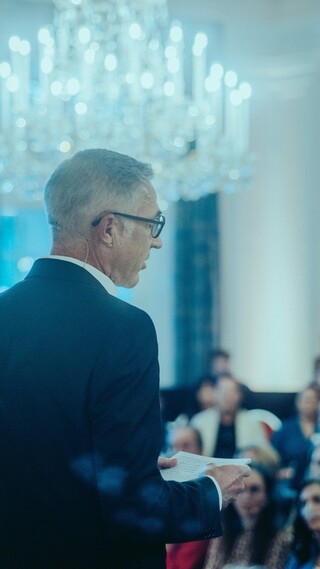 Vortragender in Anzug blickt auf sein Publikum unter einem Kronleuchter.