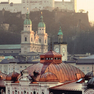 Stammhaus des Bankhaus Spängler in der Schwarzstraße 1 in 5020 Salzburg von oben. Barocke Kuppeln und eine dunkelrosa Fassade sind ersichtlich. Im Hintergrund sieht man die Festung Hohensalzburg und die Domkuppeln.