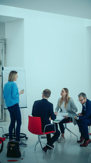 Drei Personen sitzen vor einer neben einem Flipchart stehenden Dame.