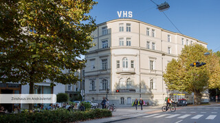 Fassade eines historischen Gebäudes mit der Aufschrift "VHS" von Bäumen umgeben.