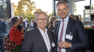 Zwei Männer in grauen Sakkos lächeln. Sie haben beide ein Glas Wasser in der Hand. Ein Mann trägt eine eckige Brille.