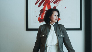 Eine Dame in Geschäftskleidung stützt sich mit den Händen auf eine Kommode und blickt zur Seite.