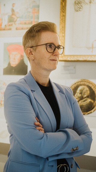 Eine Dame in Geschäftskleidung steht seitlich vor einer mit historischen Objekten behangenen Wand.