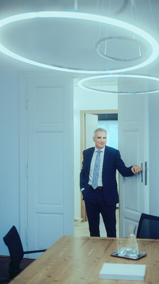 Ein Mann im Anzug, mit weißem Hemd und hellblauer Krawatte, betritt einen Besprechungsraum. Oben ist eine moderne, kreisförmige Deckenleuchte sichtbar.