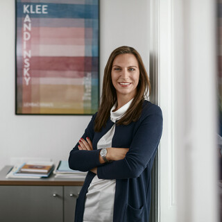 Ein Beraterin lehnt am Türrahmen in einem Bürogebäude des Bankhaus Spängler. Sie lächelt freundlich, im Hintergrund sind ein buntes Bild und ein Schrank ersichtlich. 