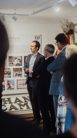 Blick über die Schulter zweier Personen auf eine Menschenansammlung vor einer Informationstafel.