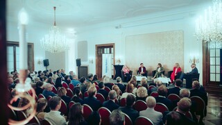 Fünf Sprecher sitzen vor einem Publikum in einem großen Saal, der mit zwei Kronleuchtern ausgestattet ist.