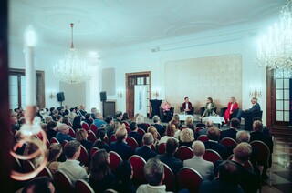 Fünf Sprecher sitzen vor einem Publikum in einem großen Saal, der mit zwei Kronleuchtern ausgestattet ist.