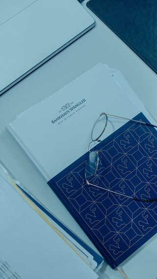 Auf dem Tisch liegt ein Notizbuch mit dem Logo vom Bankhaus Spängler. Darauf befindet sich eine Brille, links davon liegen Unterlagen mit Zahlen.