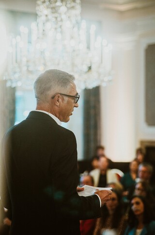 Vortragender in Anzug auf einer Bühne in einem historischen Saal.