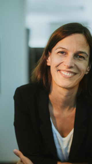 Eine lächelnde Dame in Geschäftskleidung lehnt auf einem Tisch.