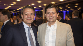 Zwei Männer lächeln. Einer trägt ein dunkles Sakko mit hellblauem Hemd und gräulicher Krawatte, der andere ein beige-farbiges Sakko und eine weißes Hemd.