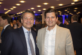 Zwei Männer lächeln. Einer trägt ein dunkles Sakko mit hellblauem Hemd und gräulicher Krawatte, der andere ein beige-farbiges Sakko und eine weißes Hemd.